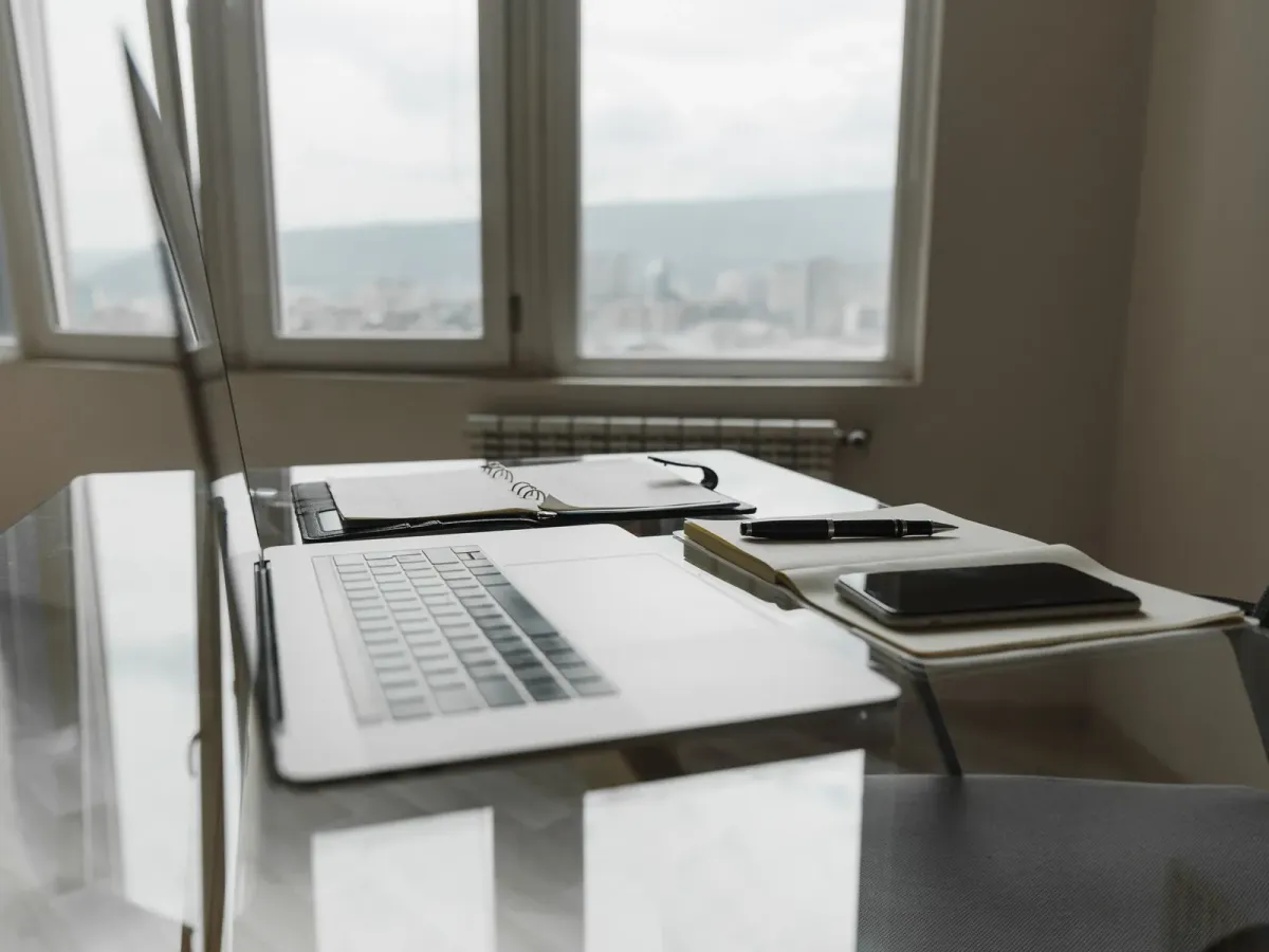 A laptop, a notebook and a pen on a glass desk by a window overlooking the city