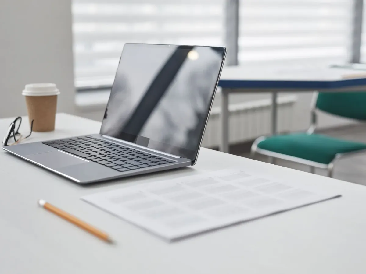 Ordinateur portable ouvert, feuille imprimée et crayon sur un bureau blanc, près d’une fenêtre