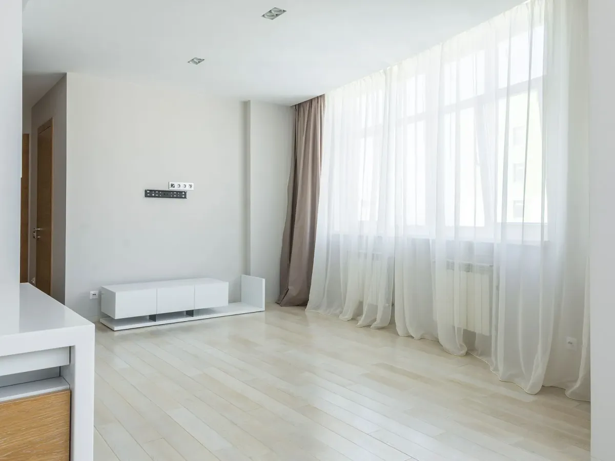 A bright, empty apartment with light wooden floors and sheer white curtains
