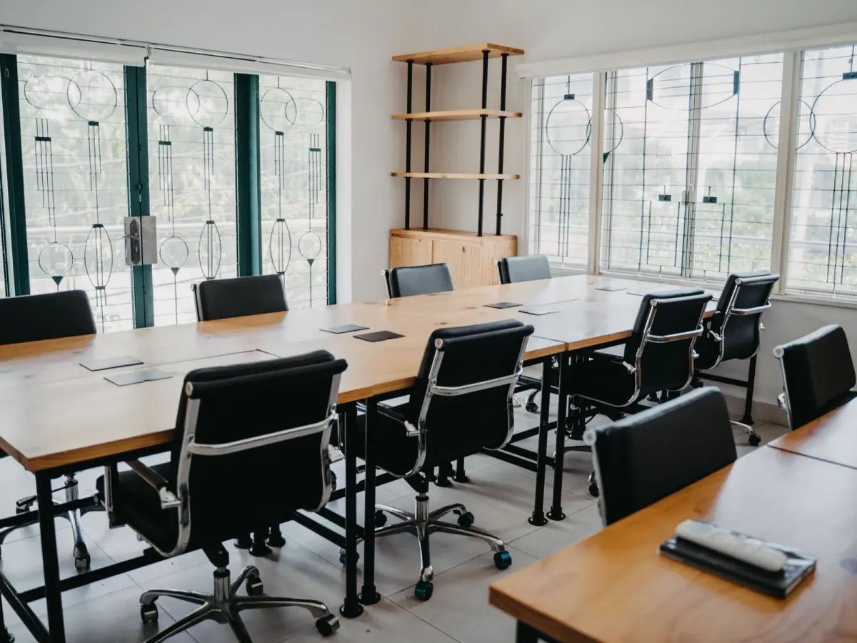 A bright meeting room with wooden tables and black chairs by tall windows