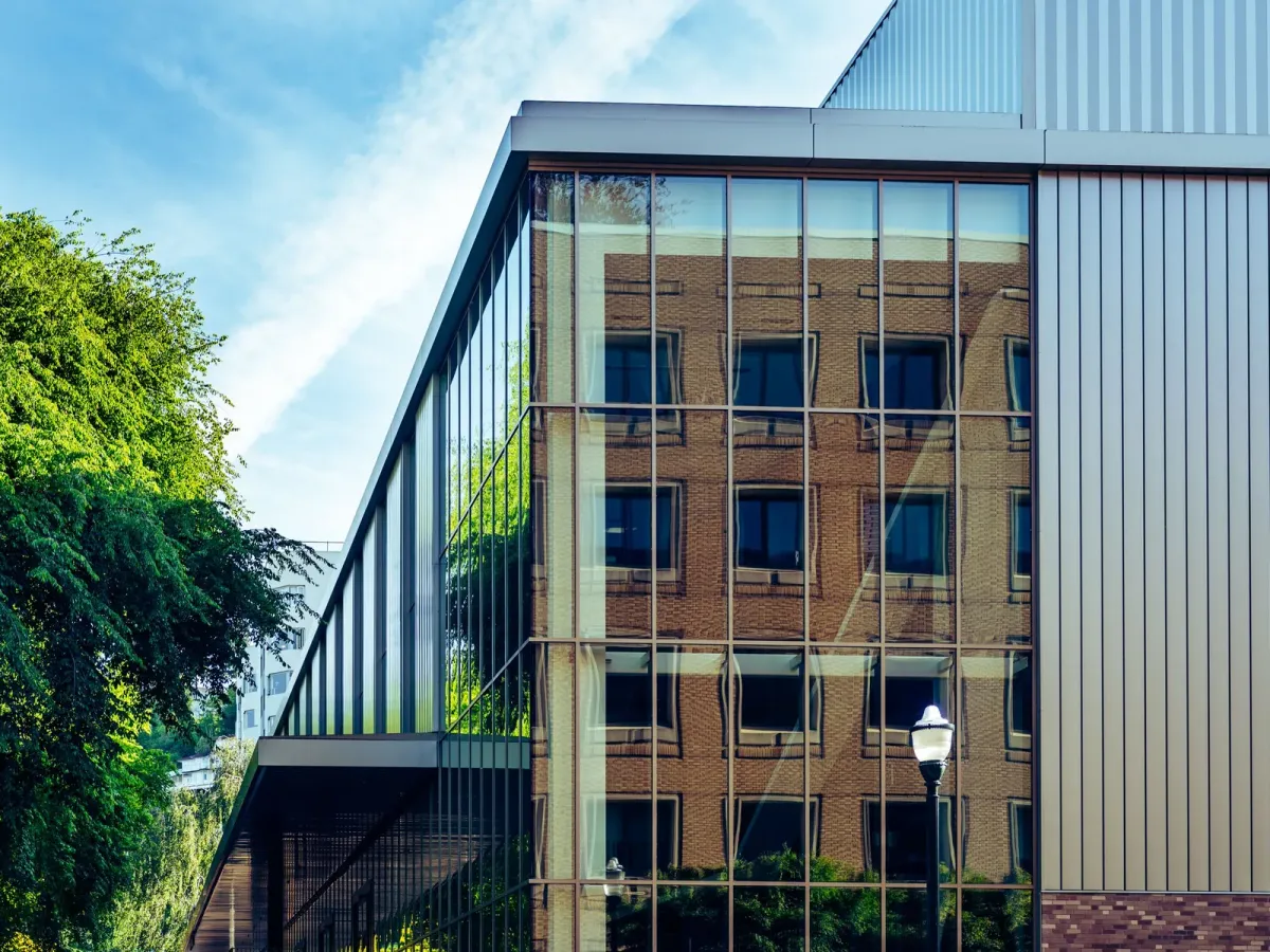 Façade vitrée d’un immeuble récent qui reflète un bâtiment de brique, près d’un arbre