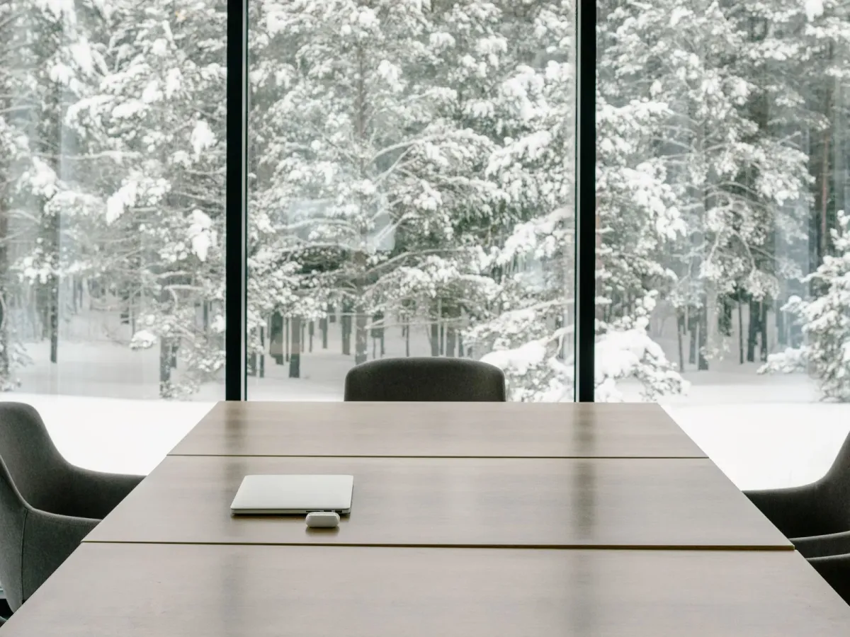 Salle de réunion vitrée avec une table en bois et des fauteuils gris, face à une forêt enneigée