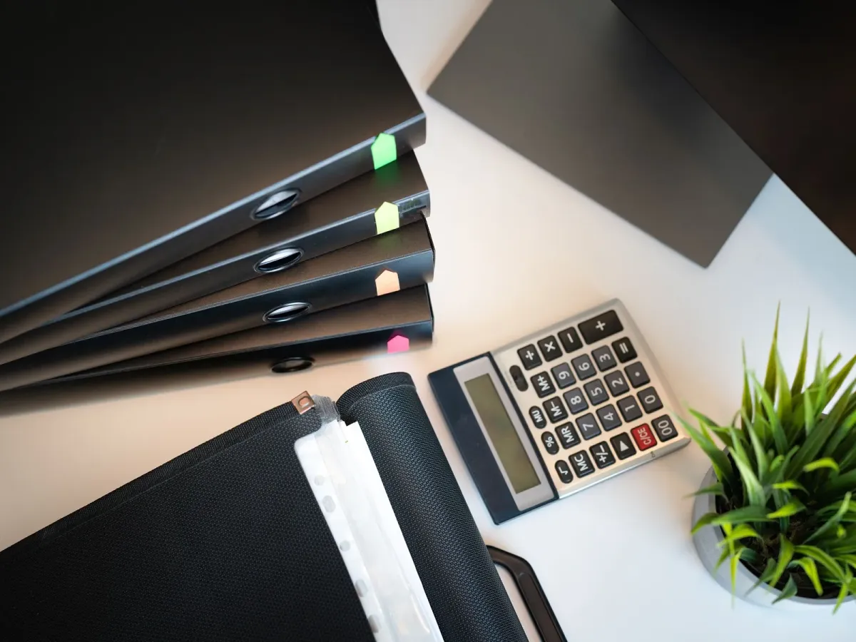 Black binders, a calculator and a small plant on a white desk, seen from above