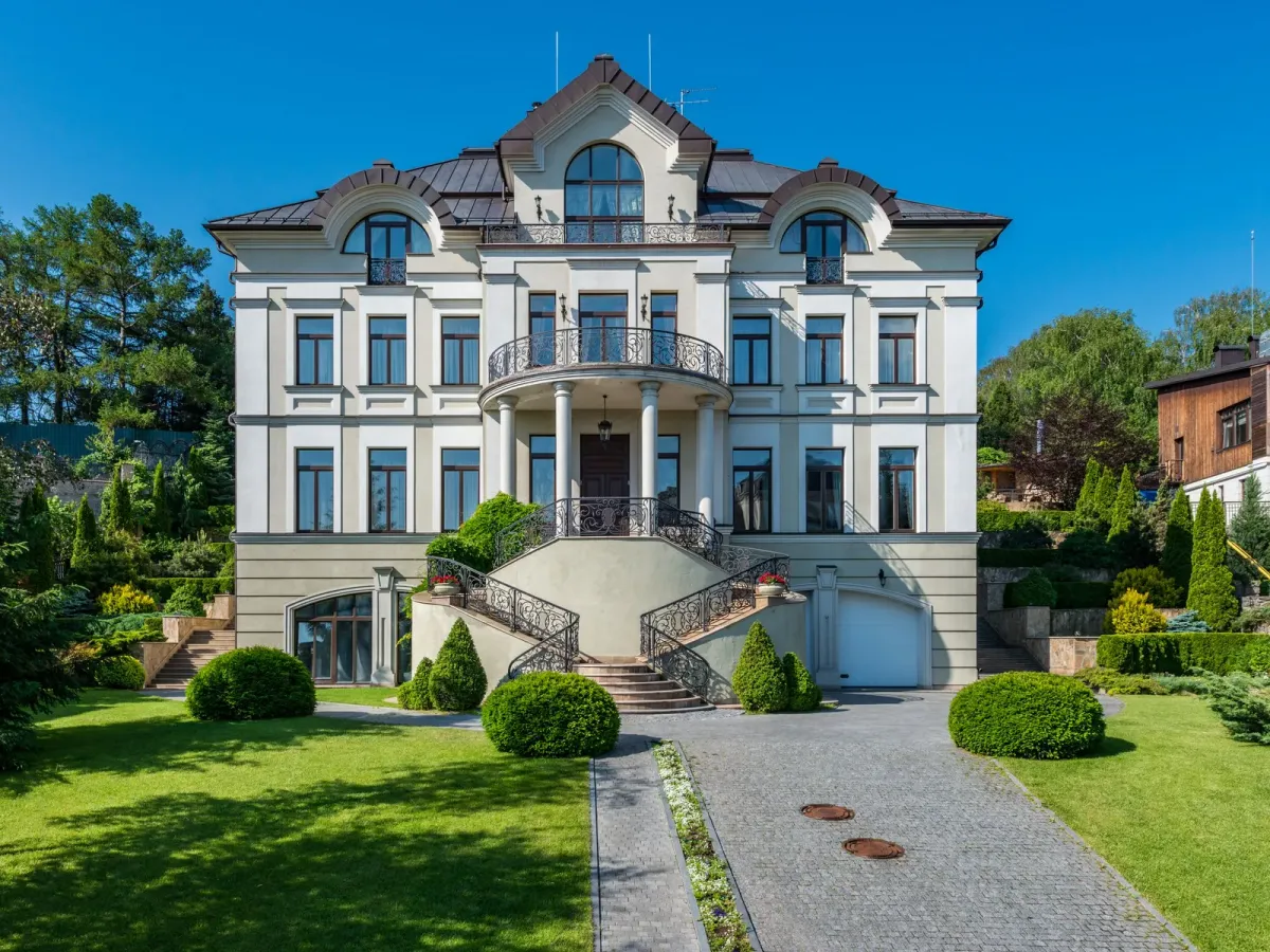 A large white villa with a central balcony and stairs, beyond a lawn and trimmed shrubs