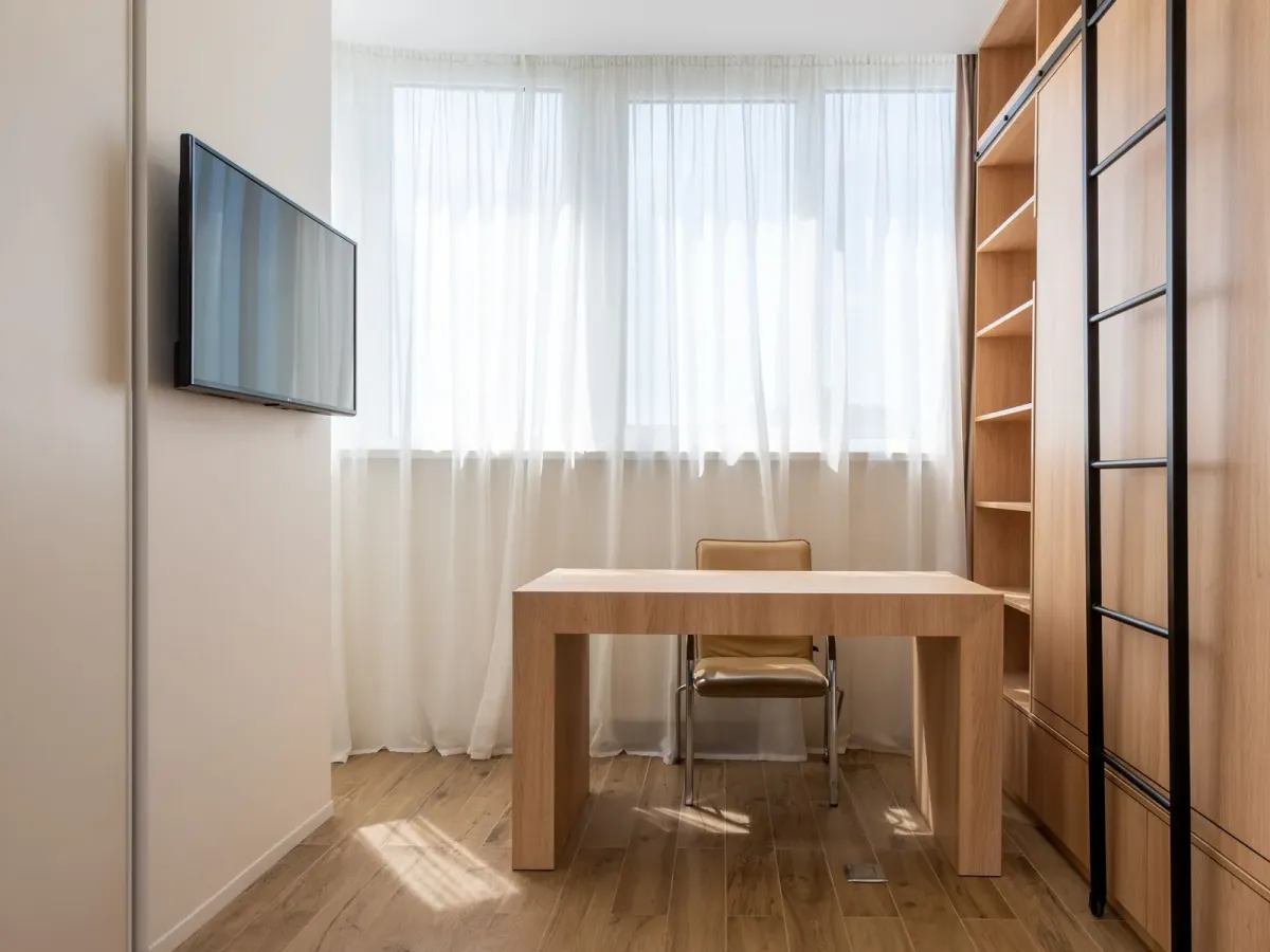 Table de travail en bois et chaise devant une fenêtre voilée, dans une pièce claire aux étagères de bois