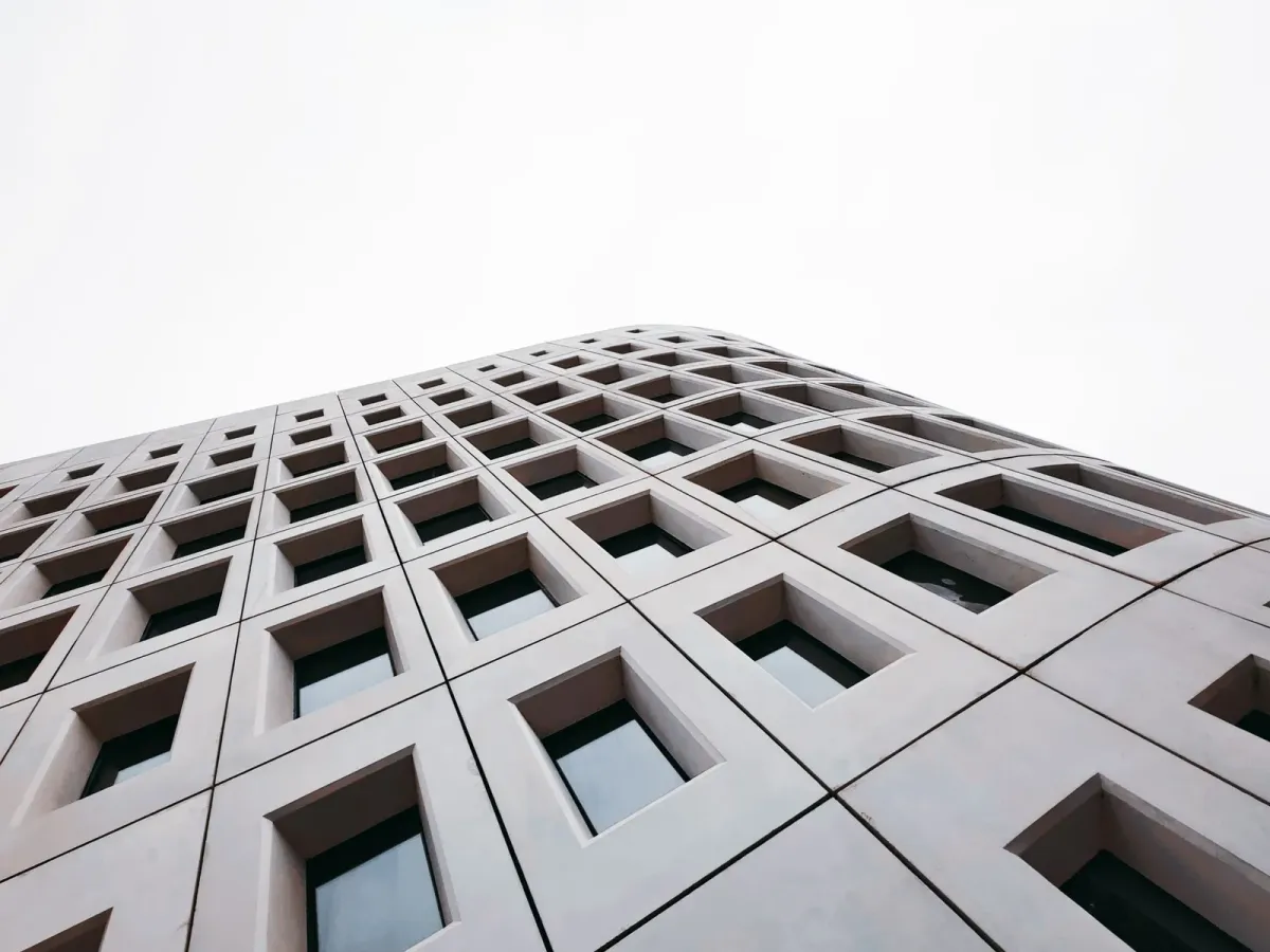 Façade de béton clair aux fenêtres carrées en retrait, vue d’en bas sous un ciel blanc