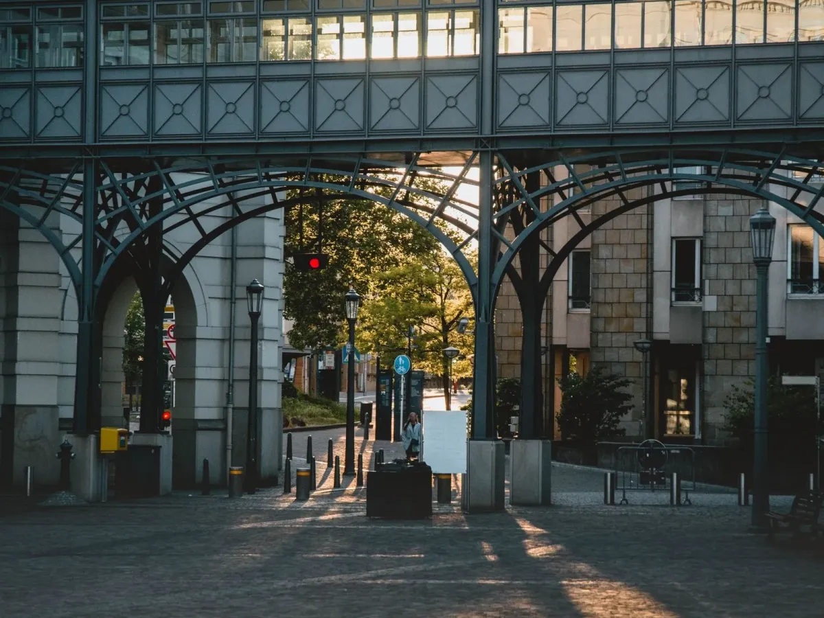 Arcades métalliques d’une passerelle traversées par la lumière du soir, au-dessus d’une place pavée